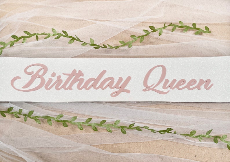 May include: A white banner with the words "Birthday Queen" written in pink cursive letters. The banner is decorated with green leafy vines.