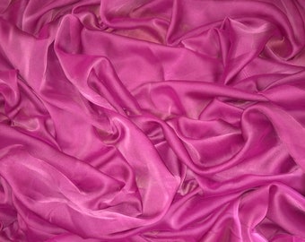 Op de afbeelding: Een close-up van een zachte, roze stof met een zijdeachtige textuur. De stof is gedrapeerd en gevouwen, wat een vloeiende en elegante uitstraling creëert.