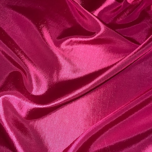 Puede incluir: Primer plano de una tela de color rosa intenso con una textura suave y sedosa. La tela está drapeada y doblada, creando líneas suaves y fluidas.
