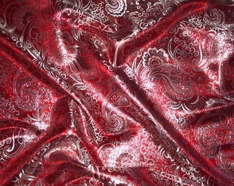 May include: A close-up of a red fabric with a silver paisley pattern. The fabric is draped, creating folds and shadows that highlight the intricate design. The shiny material reflects light, adding depth to the textile.