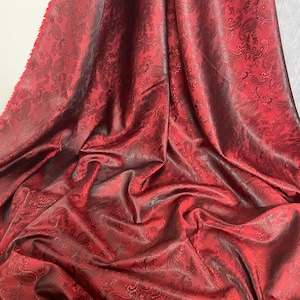 May include: Red paisley patterned fabric with a satin finish. The fabric is draped and folded, showing the texture and sheen.