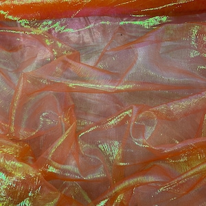 Op de afbeelding: Close-up van iriserende stof in oranje en groene tinten. Het doorschijnende materiaal is gelaagd en gevouwen, waardoor een textuureffect ontstaat. De stof heeft een iriserende kwaliteit, met kleuren die veranderen afhankelijk van het licht. Waarschijnlijk gebruikt voor decoratieve doeleinden.