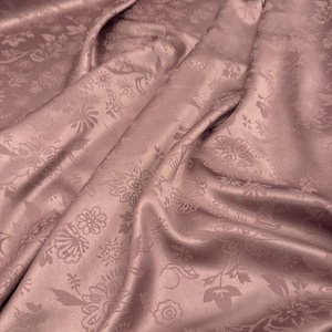 Può includere: Primo piano di un tessuto di raso rosa antico con un motivo floreale. Il tessuto è drappeggiato, mostrando pieghe e ombre. Il motivo floreale è sottilmente impresso, aggiungendo consistenza e profondità al materiale. Ideale per abbigliamento o arredamento.