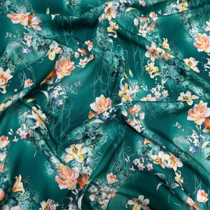 Pode incluir: Tecido verde esmeralda com um padrão floral. O tecido apresenta um design repetido de flores brancas, amarelas e cor de pêssego com folhas verdes. O tecido é drapeado, criando sombras e destaques.