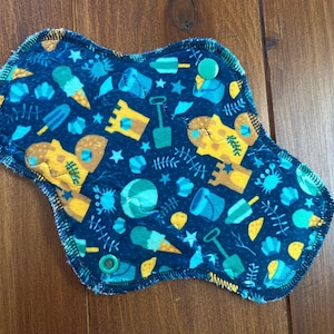 May include: A reusable cloth menstrual pad featuring a dark blue background with a repeating pattern of yellow sandcastles, blue buckets, shovels, ice cream cones, and small stars. The pad has two green plastic snap buttons for secure closure.
