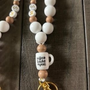 Puede incluir: Un cordón de cuentas blancas y marrones con un llavero dorado. El cordón tiene un dije con una taza de café blanca que dice "Coffee fuels teachers".