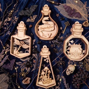 Può includere: Quattro ornamenti in legno tagliati al laser a forma di bottiglia con disegni intricati: una pergamena con il testo The Shapeshifter Brew, una chiave alata, un calderone ribollente su un fuoco e funghi, ciascuno con un tappo di sughero.