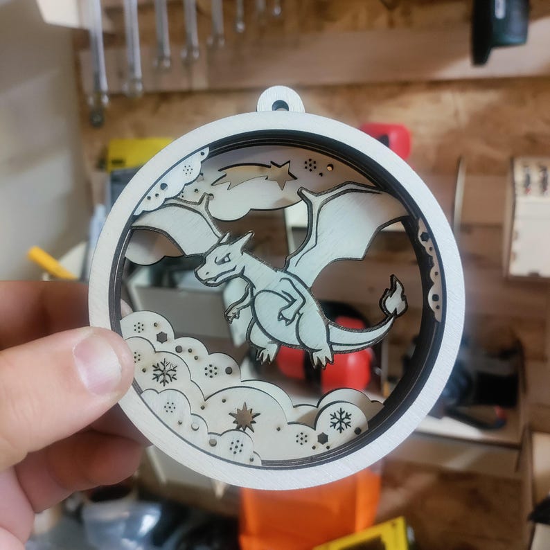 May include: A round, wooden ornament featuring a dragon design. The dragon is light gray with black outlines, set against a backdrop of clouds and snowflakes. The ornament has a circular frame with a small hole for hanging.