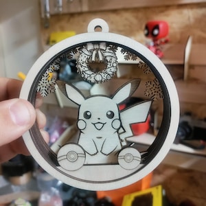 May include: Wooden Christmas ornament featuring a Pikachu design. The ornament is circular with a layered design, showcasing Pikachu, snowflakes, and a wreath. The ornament is made of light-colored wood with a loop for hanging.