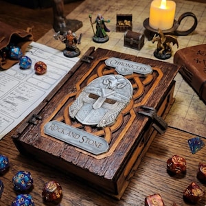 Può includere: Un libro di legno Chronicles of Rock and Stone con un design a elmo argentato. Il libro è circondato da dadi, miniature e una candela accesa. La copertina del libro presenta il testo "Chronicles of" e "Rock and Stone".