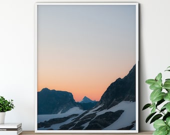 May include: A framed photograph of a mountain range with a pink and orange sunset in the background. The mountains are covered in snow and the sky is a light blue.