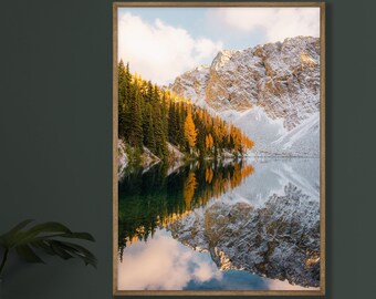 May include: A framed photograph of a mountain lake with snow-capped peaks reflected in the water. The lake is surrounded by evergreen trees, some of which have yellow leaves. The sky is a pale blue with wispy clouds.