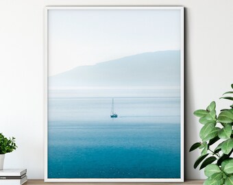 May include: A framed photograph of a sailboat on a calm body of water with a mountain range in the background. The water is a light blue color and the sky is a light gray color.
