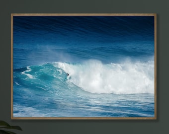 May include: A framed print of a large ocean wave, with a surfer riding the wave. The wave is a vibrant turquoise and white, set against a deep blue ocean and sky. The frame is a light brown color.