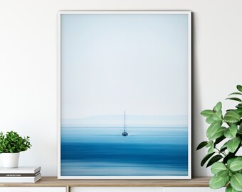 May include: A framed photograph of a sailboat on a calm, blue ocean. The sailboat is small and appears to be far away. The ocean is a light blue color and the sky is a pale blue. The photograph is in a white frame and is hanging on a white wall.