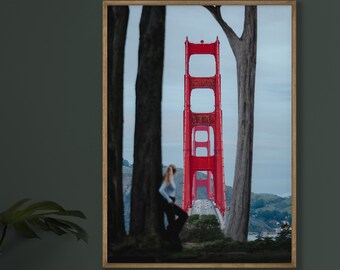 May include: A framed photograph of the Golden Gate Bridge in San Francisco, California. The bridge is red and is seen from a distance, with trees in the foreground and a person standing in the distance.