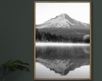 May include: A black and white photograph of a mountain reflected in a lake. The mountain is covered in snow and the lake is surrounded by trees. There is fog in the air.
