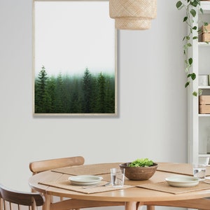May include: A framed print of a misty forest with tall evergreen trees. The print is hanging on a white wall above a round wooden dining table with four chairs. A woven pendant light hangs above the table.