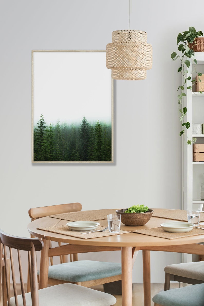May include: A framed print of a misty forest with tall evergreen trees. The print is hanging on a white wall above a round wooden dining table with four chairs. A woven pendant light hangs above the table.