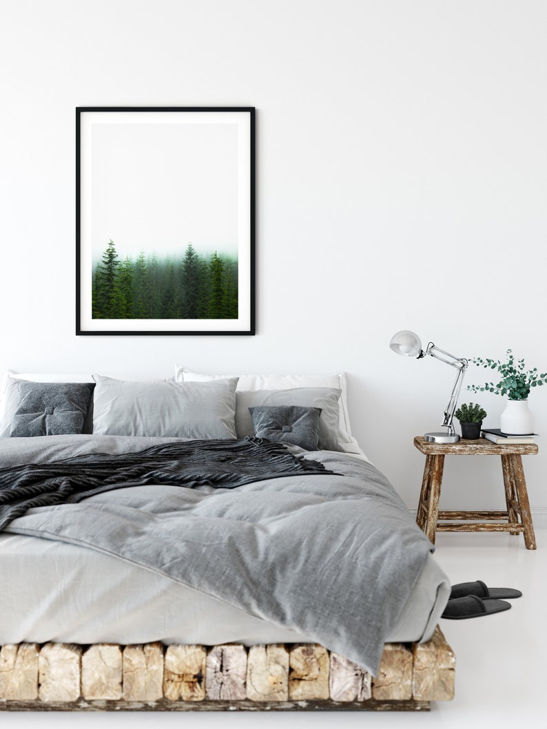 May include: A gray bed with a black throw blanket and a framed print of a foggy forest above the headboard. The bed has a wooden base and is styled with gray pillows and a wooden nightstand with a lamp and a potted plant.