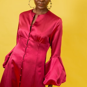 May include: A woman wearing a long-sleeved, magenta satin dress with a button-up front and large, puffy sleeves. She is wearing gold hoop earrings with green gemstones and a gold bracelet with green gemstones.