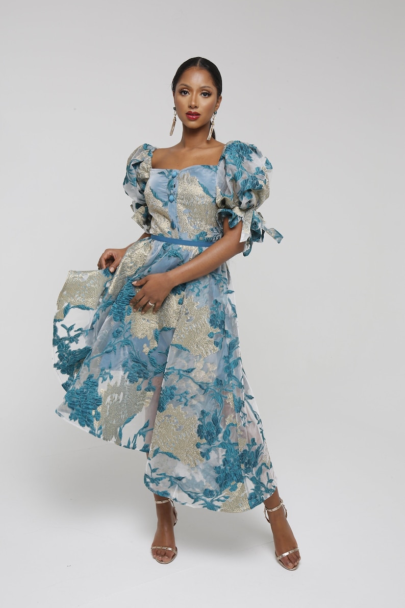 May include: A woman wearing a blue and gold floral patterned dress with puffed sleeves and a belt. She is wearing gold heeled sandals.