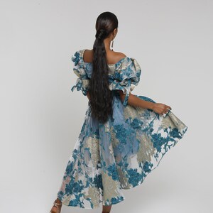 May include: A woman wearing a blue and gold floral print dress with puffed sleeves. The dress has a high low hemline and is made of a sheer fabric.