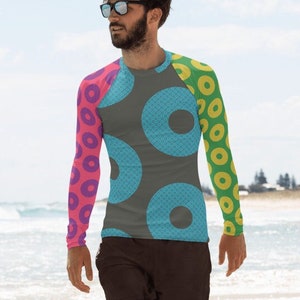 May include: A man wearing a long-sleeved shirt with a colorful donut pattern. The shirt is grey with blue, pink, and yellow donuts. The man is standing on a beach.
