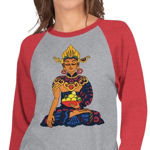 May include: A grey and red baseball tee shirt with a colorful illustration of a Buddha figure sitting in meditation holding a bowl of fruit. The Buddha is wearing a crown and ornate jewelry.