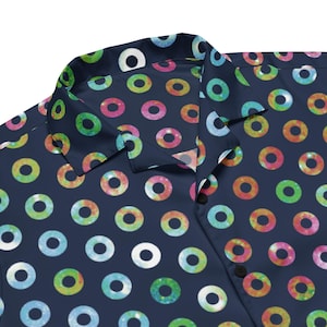 Puede incluir: Camisa abotonada azul marino con un patrón repetitivo de diseños circulares coloridos. Los círculos presentan una variedad de colores, incluyendo verde, rosa, amarillo y azul, sobre un fondo oscuro. La camisa tiene un cuello clásico y botones negros.