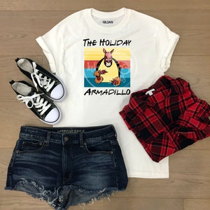Puede incluir: Camiseta blanca con un gr&aacute;fico retro de un armadillo con un gorro de Pap&aacute; Noel y el texto "The Holiday Armadillo".