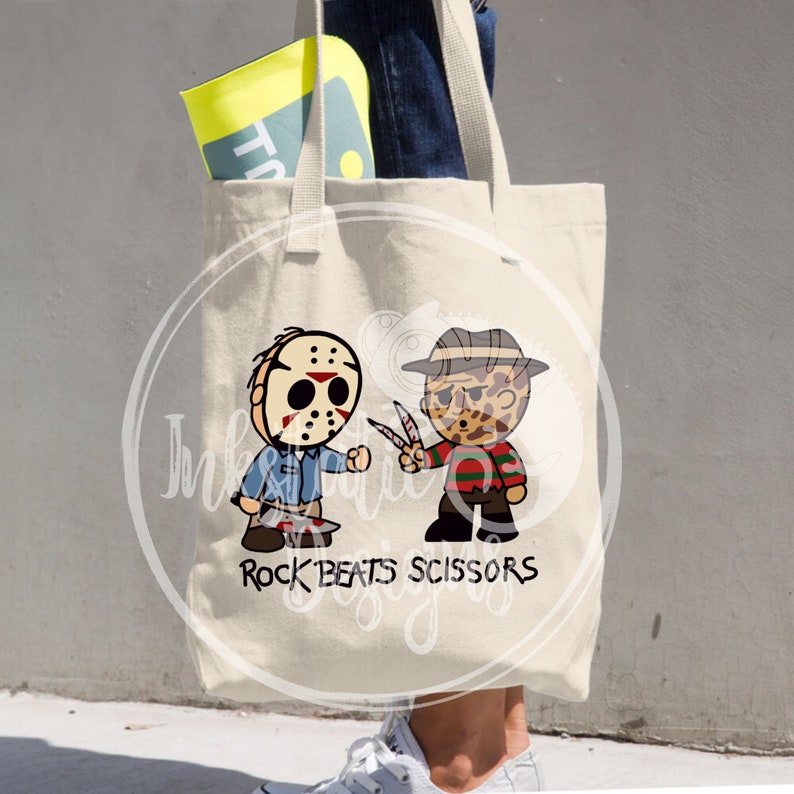 May include: Cream-colored canvas tote bag featuring a cartoon graphic of Jason Voorhees and Freddy Krueger with the text "Rock Beats Scissors". The bag has a shoulder strap and is filled with items.