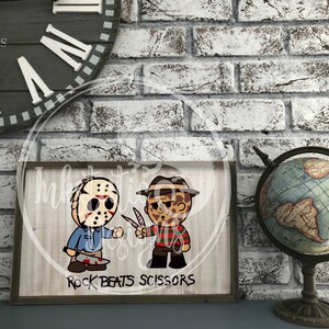 May include: A framed wooden sign featuring cartoon illustrations of Jason Voorhees and Freddy Krueger, holding a machete and knives. The sign reads "Rock Beats Scissors" in black text. The background is a light wood grain pattern.