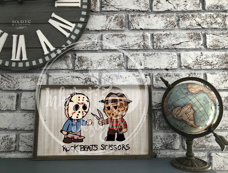 May include: A framed wooden sign featuring cartoon illustrations of Jason Voorhees and Freddy Krueger, holding a machete and knives. The sign reads "Rock Beats Scissors" in black text. The background is a light wood grain pattern.