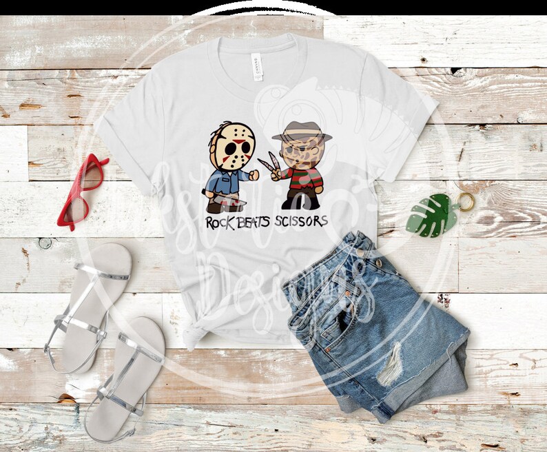 May include: White t-shirt featuring cartoon illustrations of Jason Voorhees and Freddy Krueger, with the text "Rock Beats Scissors". The shirt is paired with denim shorts, silver sandals, and red sunglasses.