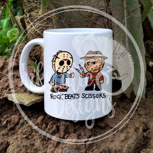 May include: White ceramic mug featuring cartoon illustrations of Jason Voorhees and Freddy Krueger. The text "Rock Beats Scissors" is printed below the characters. The mug has a handle and is set against a natural background.