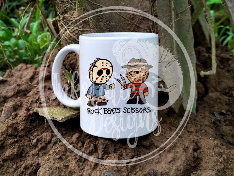 May include: White ceramic mug featuring cartoon illustrations of Jason Voorhees and Freddy Krueger. The text "Rock Beats Scissors" is printed below the characters. The mug has a handle and is set against a natural background.