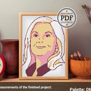 Op de afbeelding: Een ingelijst kruissteekpatroon van een vrouw met blond haar. Het patroon is in tinten roze, geel en bruin. Het afgewerkte project meet 7,4 inch bij 9,8 inch of 18,7 centimeter bij 24,8 centimeter op 16-counts Aida-stof. Het patroon gebruikt 5 DMC-garens.