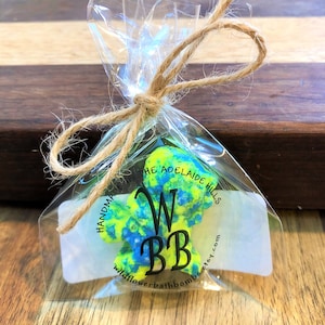 May include: A bright neon yellow and blue speckled bath bomb packaged in a clear plastic bag tied with a brown twine bow. A white label is attached to the front with the black text HANDMADE IN THE ADELAIDE HILLS WBB wildflowerbathbombs.etsy.com.