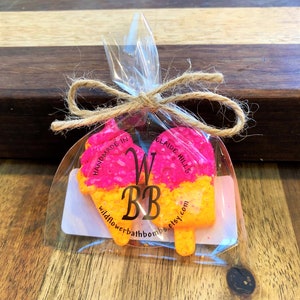 May include: Two bright pink and orange bath bombs shaped like an ice cream cone and a popsicle, packaged in a clear plastic bag tied with a brown twine bow. The label features the text WBB, HANDMADE IN ADELAIDE HILLS, and wildflowerbathbombs.etsy.com.