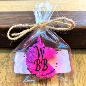 May include: A bright pink and purple unicorn-shaped bath bomb packaged in a clear plastic bag tied with a tan twine bow. A circular sticker on the front features the black text WBB, HANDMADE IN THE ADELAIDE HILLS, and wildflowerbathbombs.etsy.com.