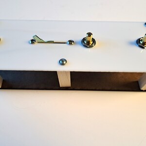 May include: A white rectangular electronic device with a wooden base. It features a toggle switch, two gold-colored knobs, and a jack. The device is secured with silver screws.