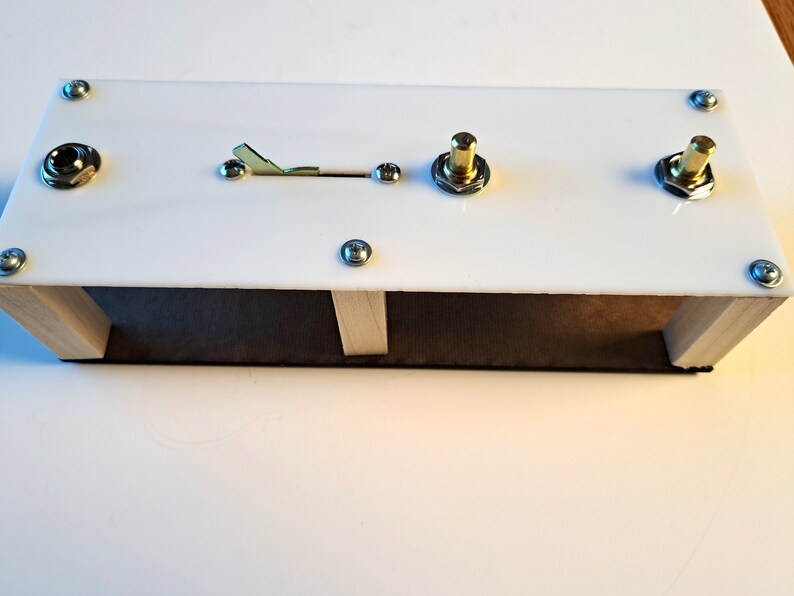 May include: A white rectangular electronic device with a wooden base. It features a toggle switch, two gold-colored knobs, and a jack. The device is secured with silver screws.