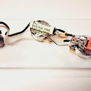 May include: A guitar wiring harness featuring a vintage tone capacitor. The components include silver potentiometers, an orange switch, and wires in black, white, and yellow. The capacitor is labelled "Jupiter Vintage Tone".