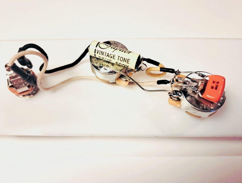 May include: A guitar wiring harness featuring a vintage tone capacitor. The components include silver potentiometers, an orange switch, and wires in black, white, and yellow. The capacitor is labelled "Jupiter Vintage Tone".