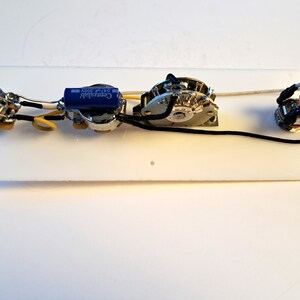 May include: Guitar electronics components, including potentiometers, capacitors, and a switch, are mounted on a white rectangular plate. The components are connected with black and yellow wires. An orange capacitor is visible.