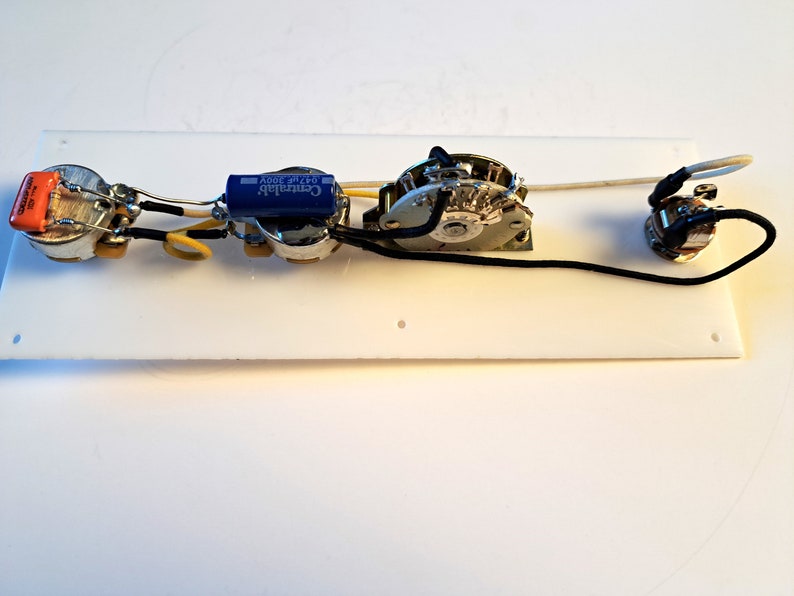 May include: Guitar electronics components, including potentiometers, capacitors, and a switch, are mounted on a white rectangular plate. The components are connected with black and yellow wires. An orange capacitor is visible.