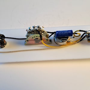 May include: A white rectangular guitar control plate with various electronic components. These include a multi-position switch, potentiometers, capacitors, and wiring. The components are connected with black, yellow, and cream-colored wires.
