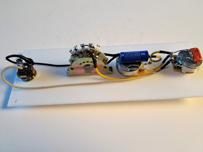 May include: A white rectangular guitar control plate with various electronic components. These include a multi-position switch, potentiometers, capacitors, and wiring. The components are connected with black, yellow, and cream-colored wires.