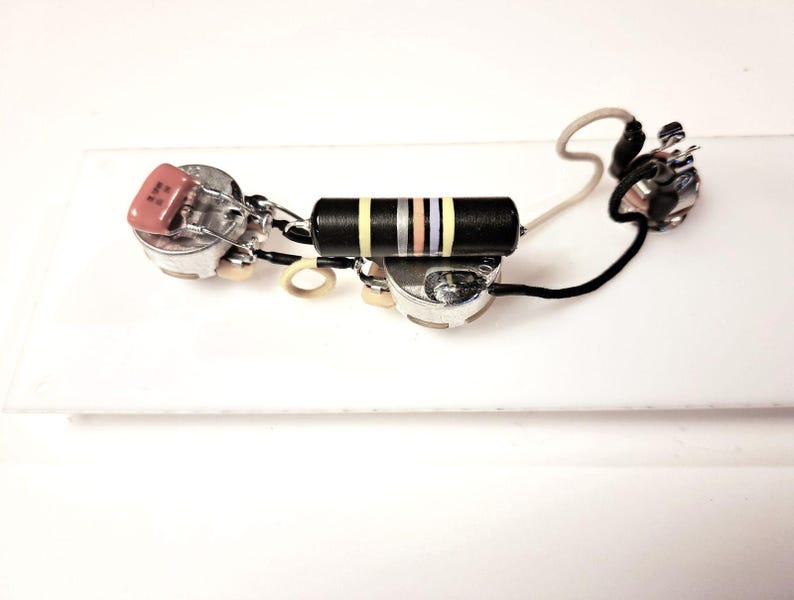 Puede incluir: Un arn&eacute;s de cableado de guitarra con varios componentes, incluyendo potenci&oacute;metros, un condensador y una resistencia con rayas amarillas, moradas y blancas. Cables negros y blancos conectan las piezas.